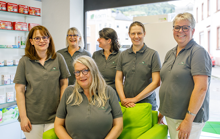 Das Team der Martinus Apotheke in Trier steht un einen grünen Sessel in der Apotheke, auf dem Sessel sitzt eine Apothekerin.