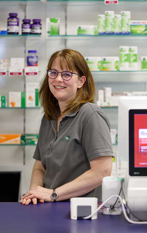 Apothekerin der Martinus Apotheke steht hinter dem Tresen vor dem Medikamentenregal und lächelt in die Kamera.
