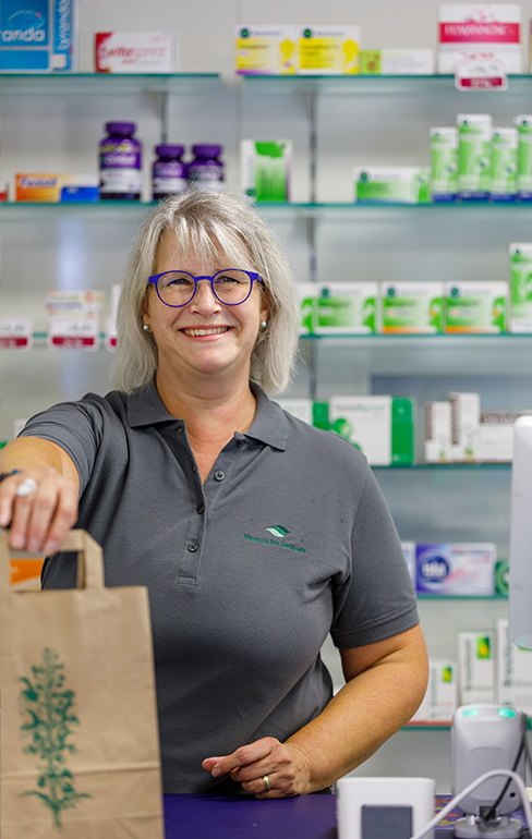 Apothekerin der Martinus Apotheke steht mit einer Tüte, hinter dem Tresen, vor dem Medikamentenregal.