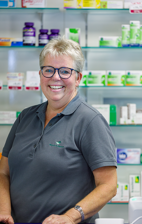 Apothekerin der Martinus Apotheke steht hinter dem Tresen vor dem Medikamentenregal und lacht in die Kamera.