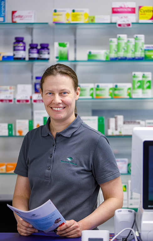 Apothekerin der Martinus Apotheke steht mit einer Broschüre, hinter dem Tresen, vor dem Medikamentenregal.