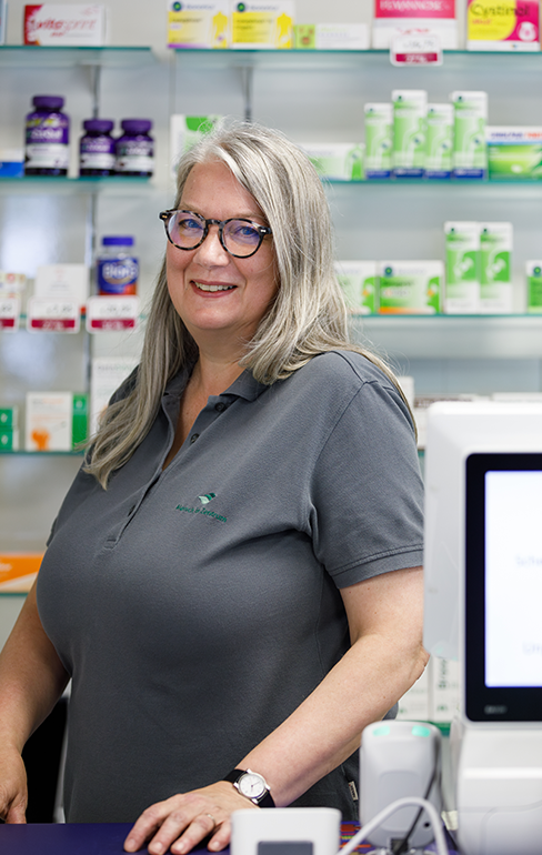 Apothekerin der Martinus Apotheke steht hinter dem Tresen vor dem Medikamentenregal und schaut nach vorne zur Kamera.