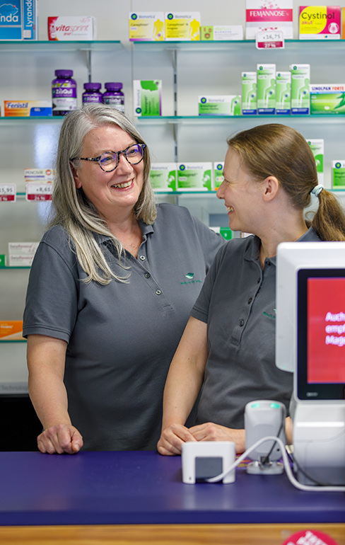 Zwei Apothekerin der Martinus Apotheke stehen hinter dem Tresen, vor dem Medikamentenregal und sprechen miteinander.