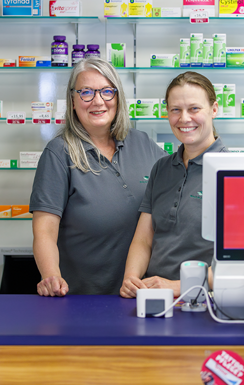 Zwei Apothekerin der Martinus Apotheke stehen hinter dem Tresen, vor dem Medikamentenregal.