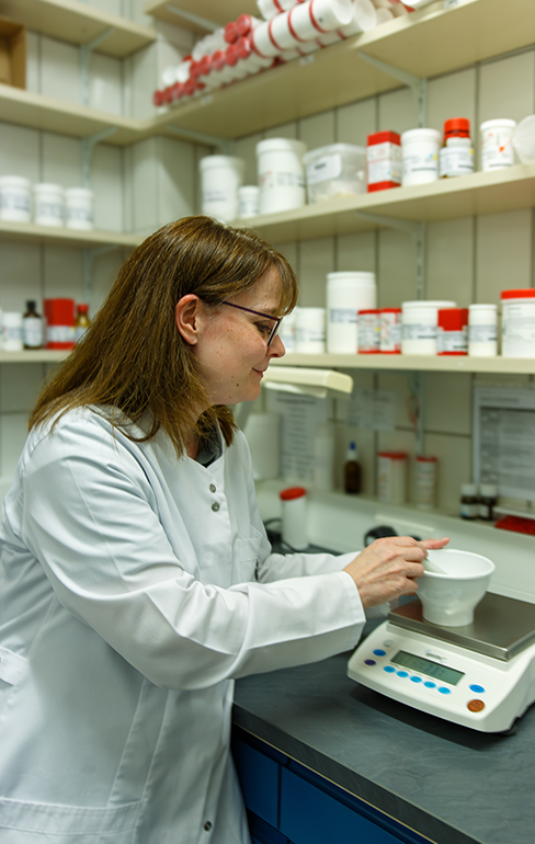 Apothekerin steht im Labor und erstellt eine Rezeptur, im Hintergrund sieht man ein Medikamentenregal.