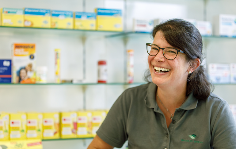 Apothekerin der Martinus Apotheke steht lachend vor dem Medikamentenregal in der Apotheke.