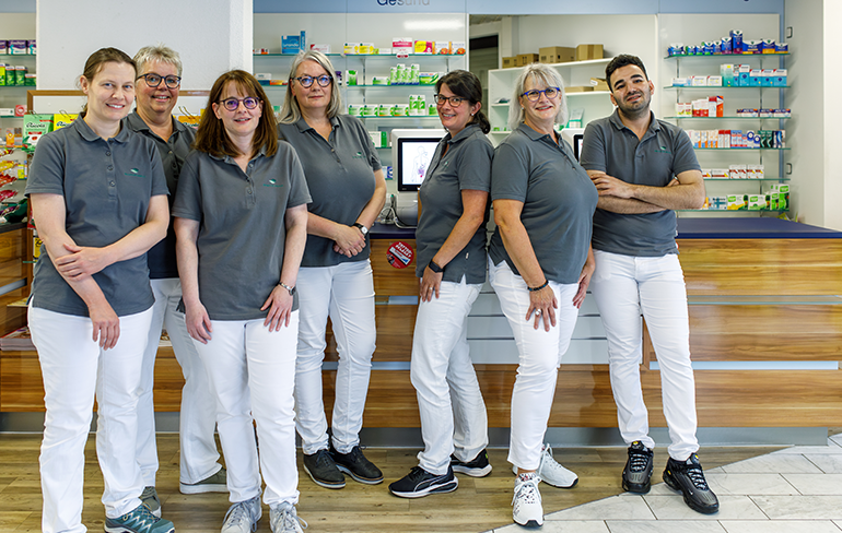 Das Apothekenteam steht vor dem Tresen in der Martinus Apotheke, im Hintergrund sieht man ein Medikamentenregal.