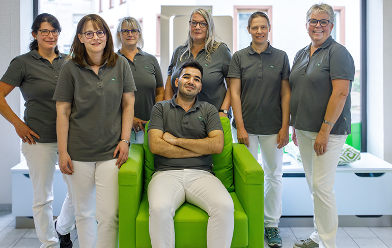 Das Team der Martinus Apotheke in Trier steht un den sitzenden Inhaber der Apotheke, der in einem grünen Sessel von dem Fenster sitzt.